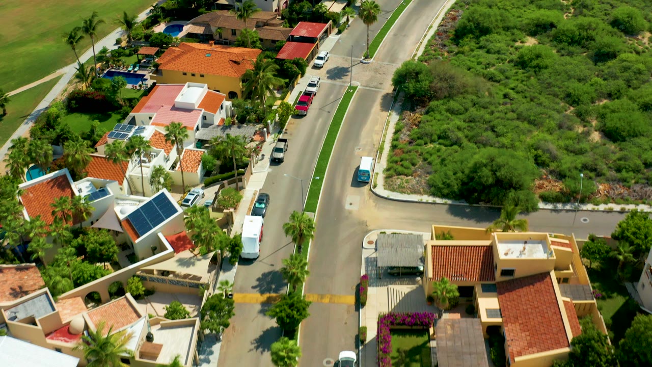 Aerial frame from San Jose Del Cabo showing a curved road, orange and white roofed homes, palm trees, parked vehicles, and dense green brush with no visible people.