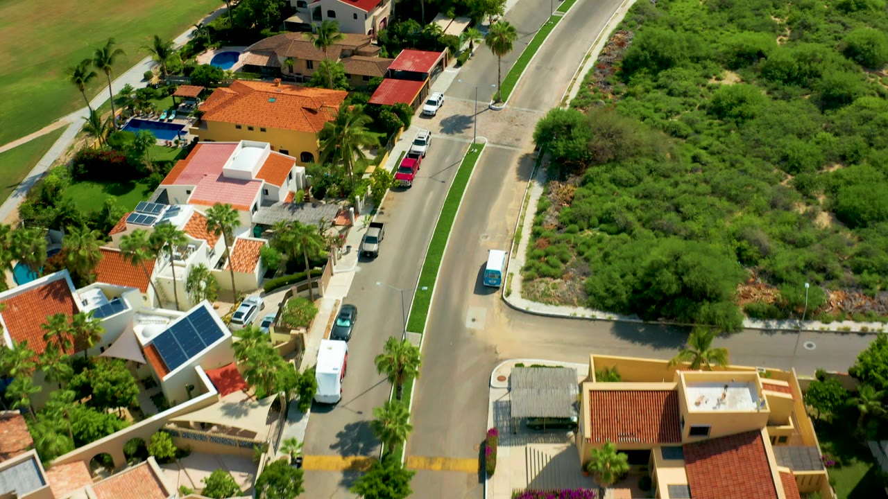 San Jose Del Cabo aerial frame showing villas with red and white roofs beside a paved road, palm trees, parked vehicles, and a large green brush field with no visible people.