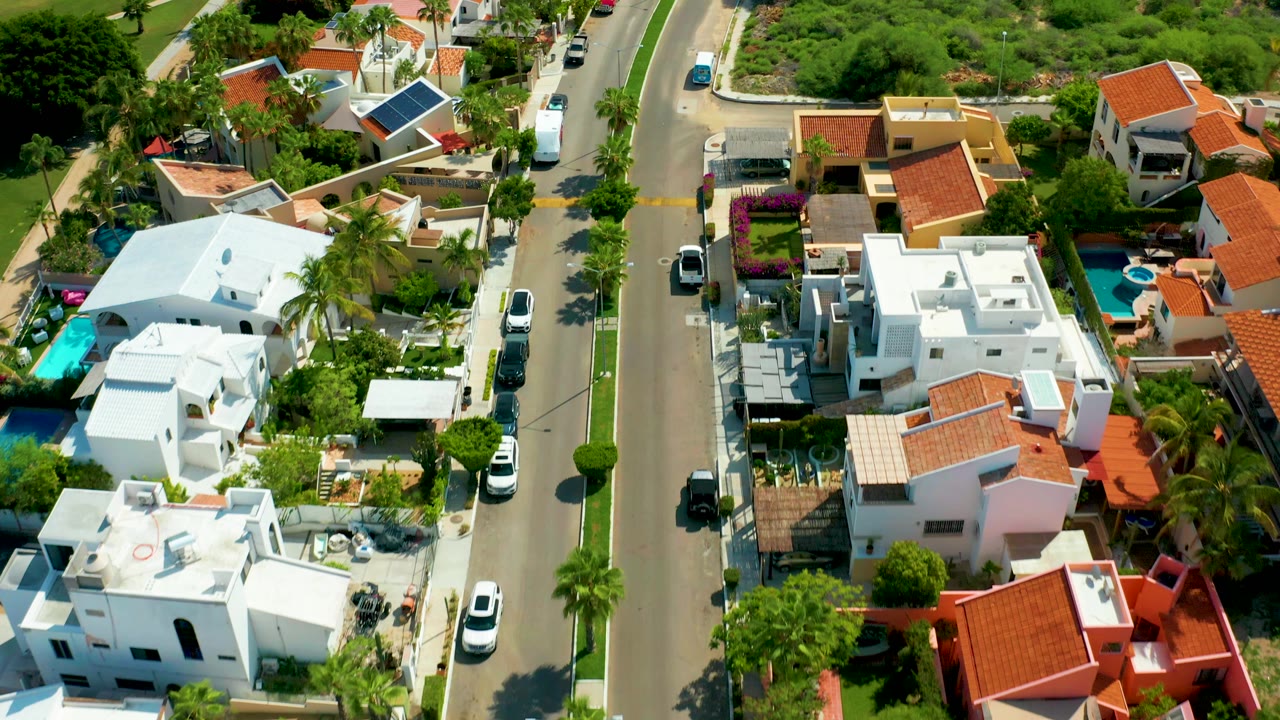 Aerial view of a San Jose Del Cabo residential street with white homes, terracotta roofs, palm trees, parked vehicles, pools, and no visible people.