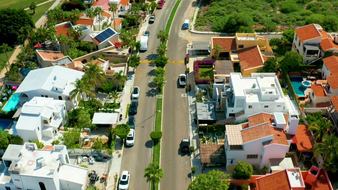 Aerial view in San Jose Del Cabo showing a curved street beside dense green open land, white and tan homes, red roofs, curbside vehicles, and no visible people.