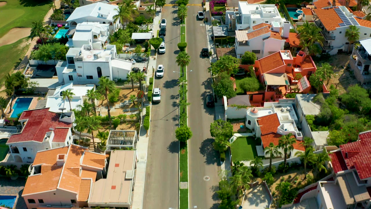 Aerial frame over San Jose Del Cabo homes with a straight paved street, white stucco buildings, red tile roofs, palms, parked cars, green yards, and no visible people.
