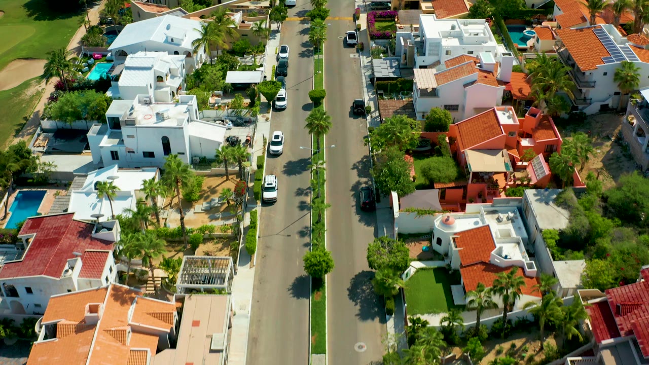 San Jose Del Cabo aerial frame showing a wider spread of white residences, red tile roofs, blue pools, palm trees, a paved street, and no visible people.