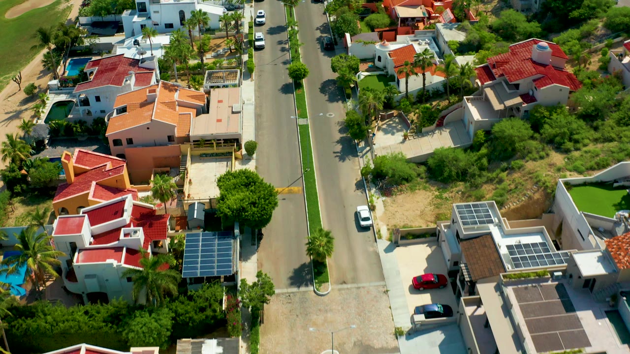 Opening aerial frame over San Jose del Cabo homes with red and orange roofs, a vertical street, solar panels, palms, parked cars, and no visible people.