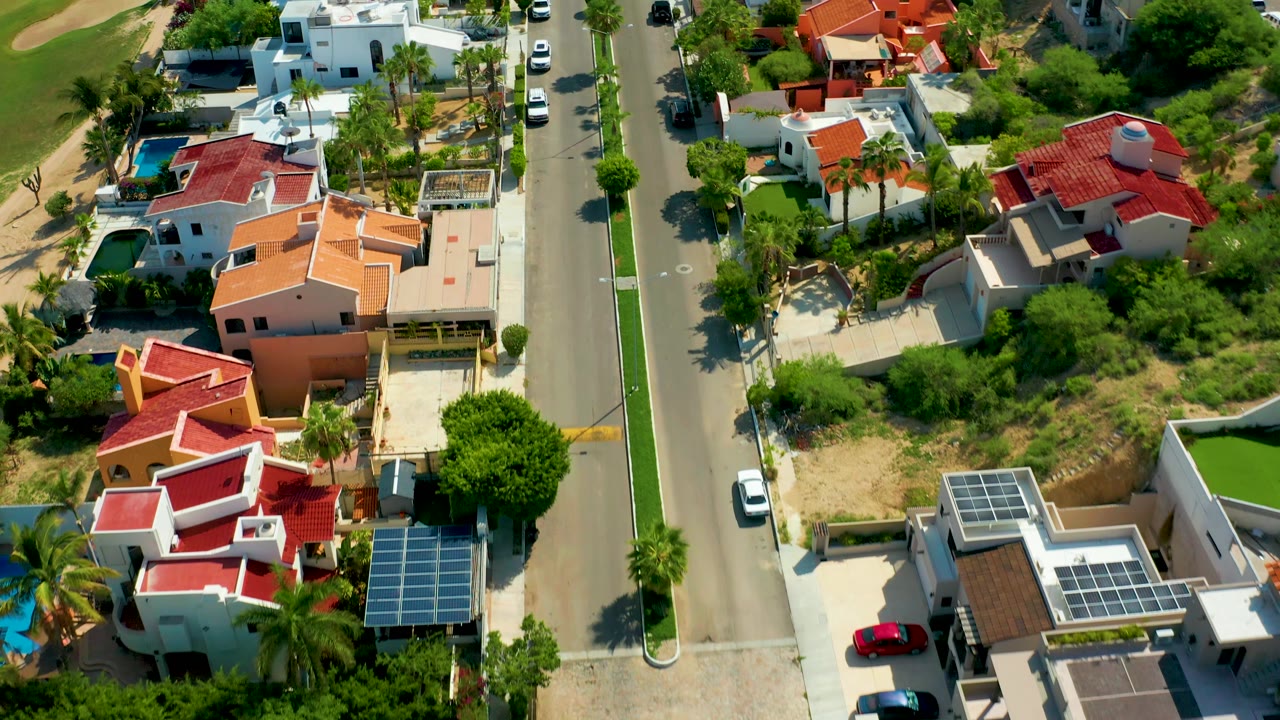 Mid-clip aerial over San Jose del Cabo residences with a white car on the street, red tile roofs, rooftop solar arrays, palms, mixed green and dry ground, and no visible people.