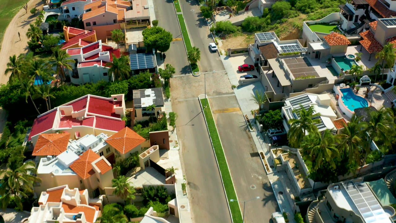 Angled aerial view over San Jose Del Cabo homes with red and orange roofs, palm trees, a straight residential street, parked cars, pools, and no visible people.