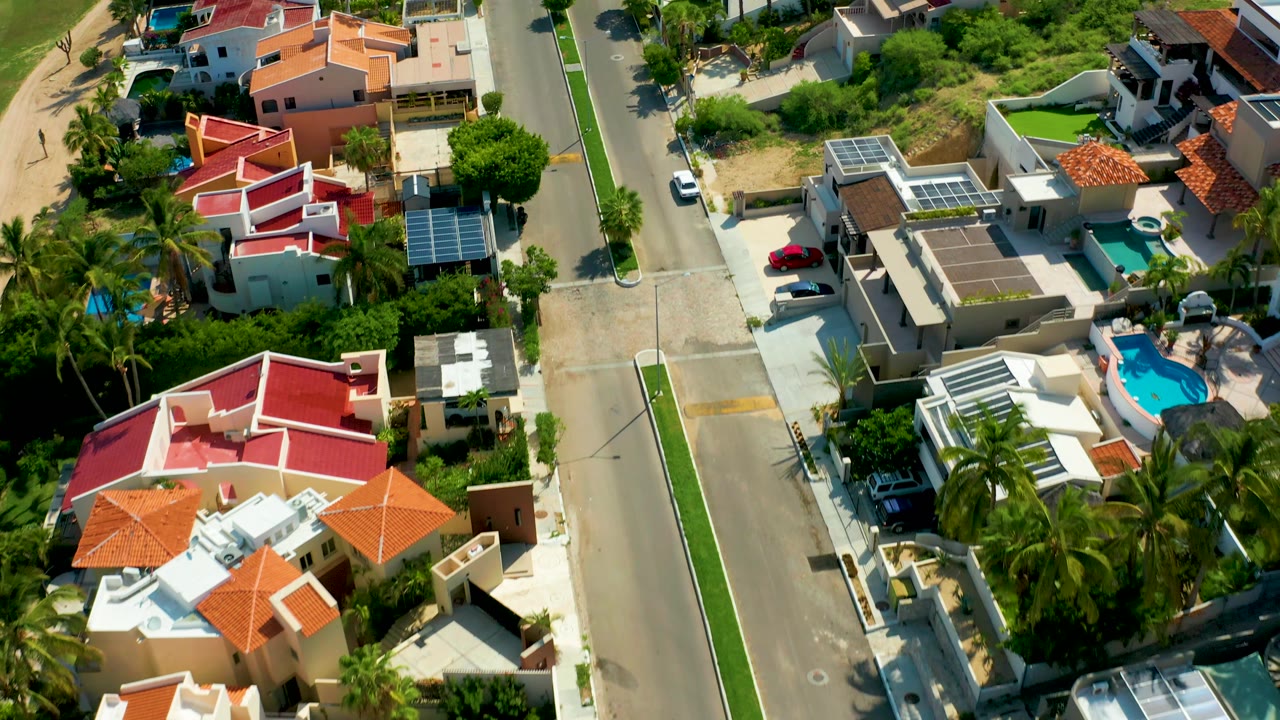 Higher angled view of a San Jose Del Cabo residential street with the white car farther along the road, roof terraces, palm clusters, pools, and no visible people.