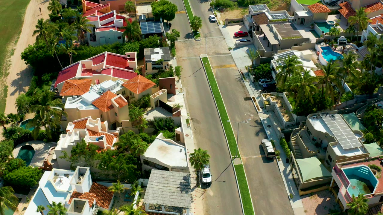 Overhead view in San Jose Del Cabo showing red and white roofed homes, palms, small pools, a paved street, and no visible people at the start of the clip.