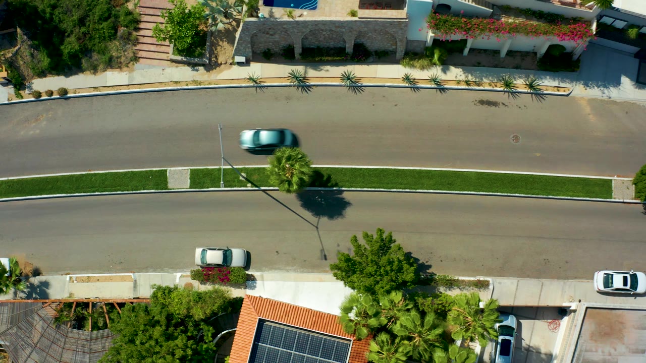 Aerial frame from San Jose Del Cabo with the blue car advancing along the upper lane, white parked cars below, landscaped yards, tiled roofs, and no visible people.