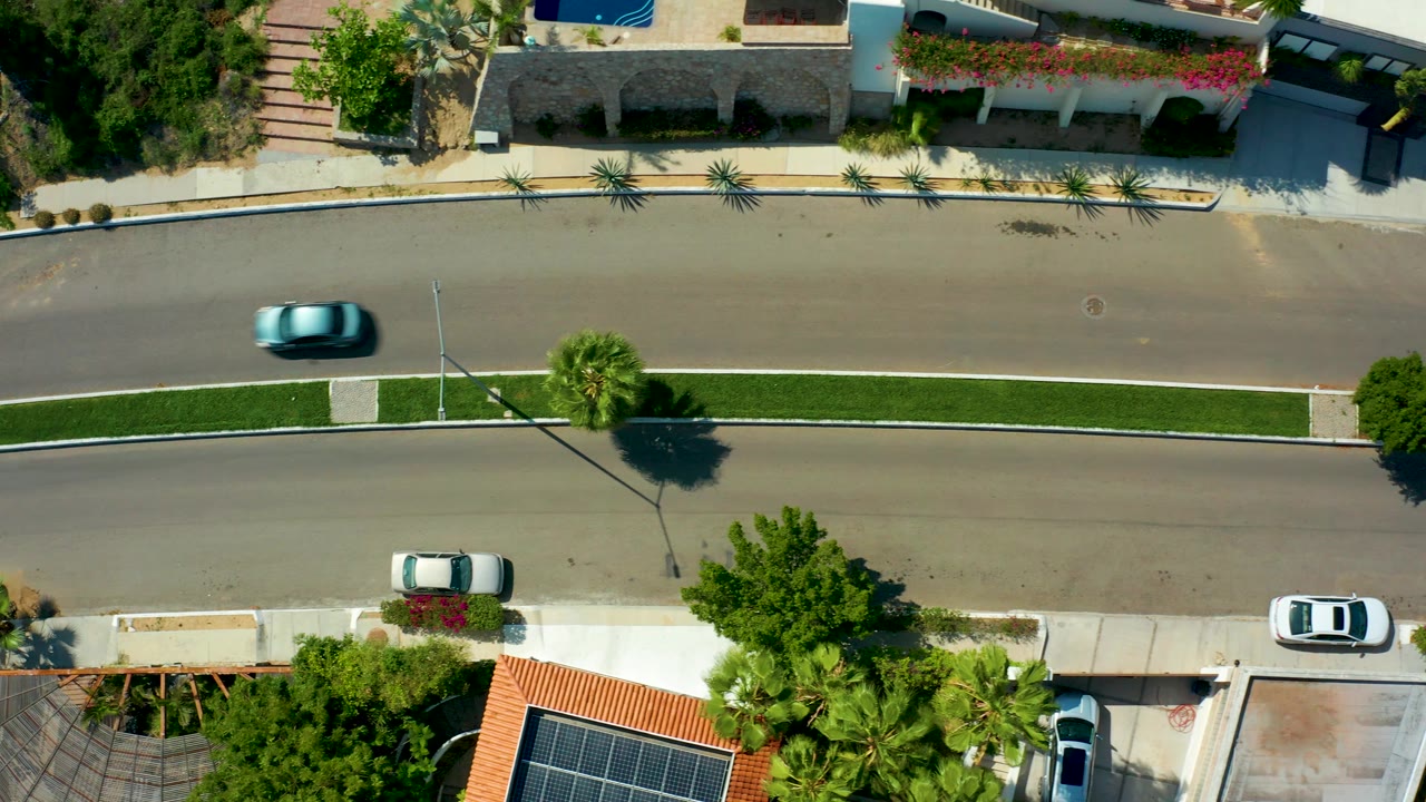 Top-down San Jose Del Cabo residential street frame with the blue car near the middle-right upper road, parked vehicles, palm trees, sidewalks, and no visible people.