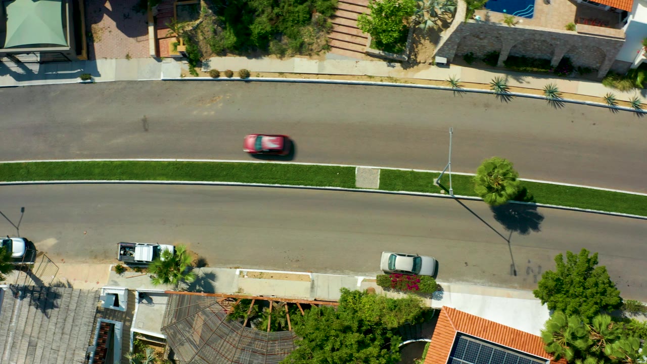 Straight-down view of a San Jose del Cabo residential road with a red car on the upper lane, parked white vehicles, sidewalks, palms, tile roofs, and no visible people.