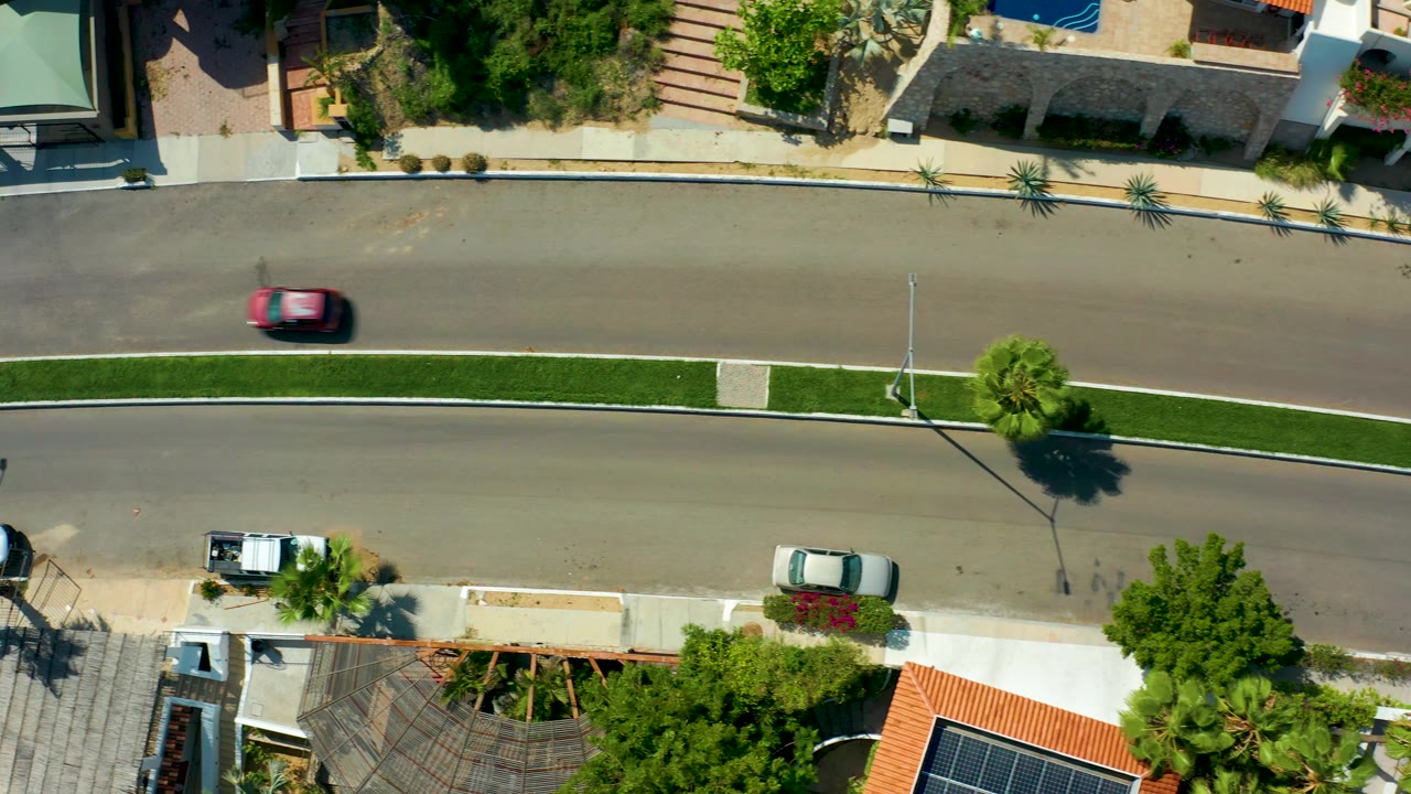 Overhead San Jose del Cabo street frame with the red car near the right side of the upper lane, a narrow green median, parked vehicles, palms, and no visible people.