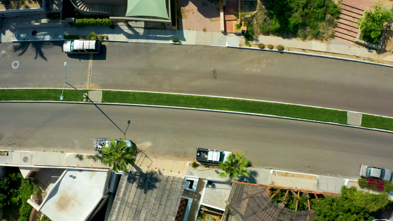 Top-down aerial frame in San Jose Del Cabo showing a white van on the upper road, houses and landscaping along both edges, a central grass strip, and no visible people.