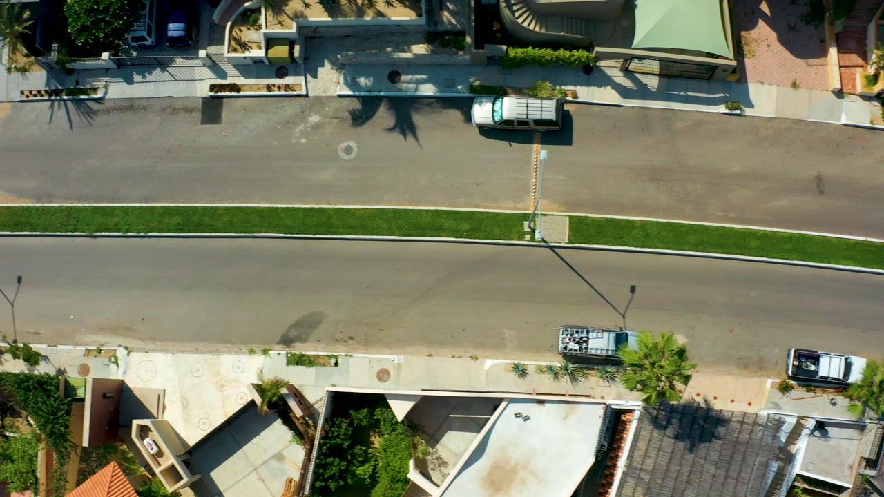 Overhead frame in San Jose del Cabo showing a white van on the upper residential road, sidewalks, homes, palm shadows, a grassy median, and no visible people.