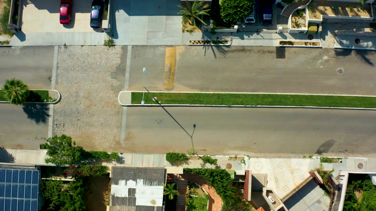 Top-down frame from San Jose del Cabo with the two-lane road crossing the image, a grass median below it, parked vehicles near the top, roof structures at the bottom, and no visible people.