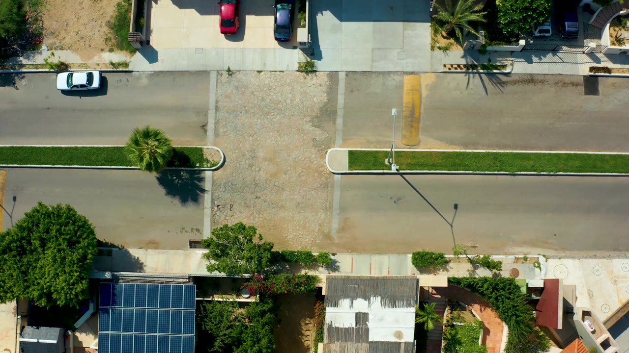 Overhead San Jose del Cabo street view with the white car near the right edge, green median strips, palms, parked cars, house roofs, solar panels, and no visible people.