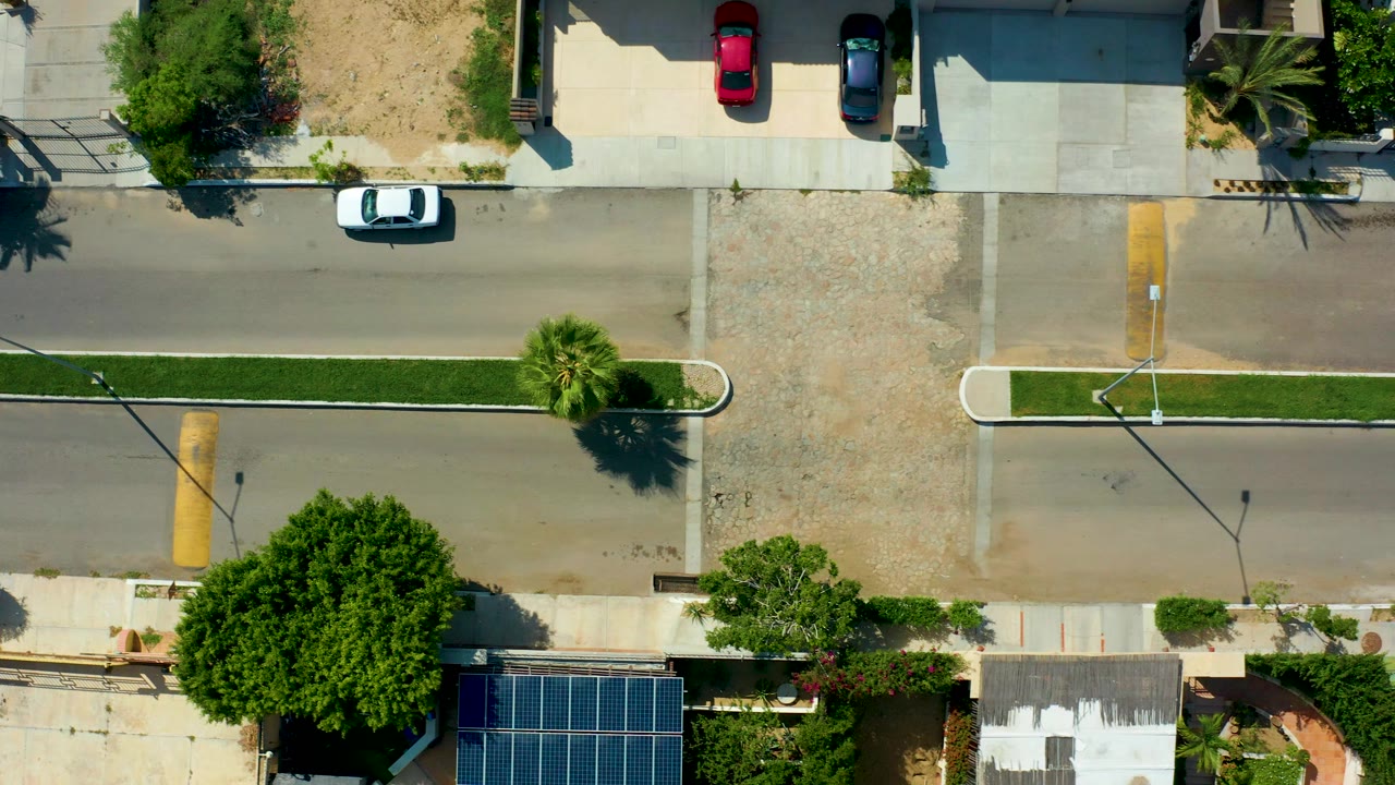 Opening overhead frame in San Jose Del Cabo showing a white car on the left side of a sunlit residential road, with a green median, sidewalks, trees, rooftops, solar panels, and no visible people.
