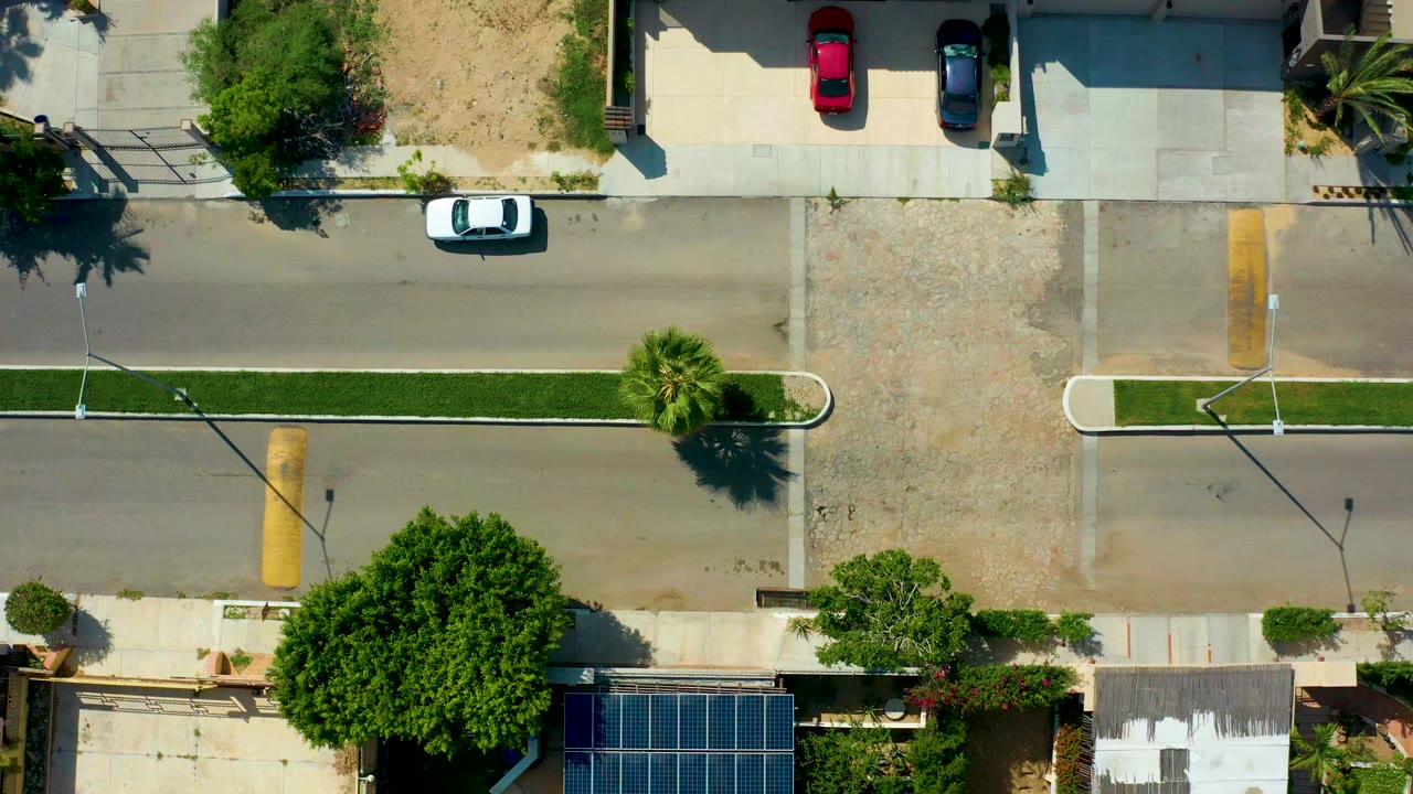 Opening overhead frame of a San Jose del Cabo residential street with parked cars, private homes, a grassy median, solar panels, trees, and no visible people.