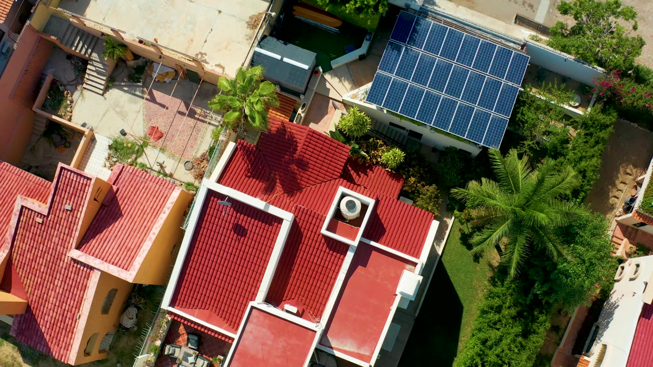 Top-down San Jose Del Cabo frame with the red roof filling the lower center, a blue solar panel array near the upper right, palms, dense greenery, and no visible people.