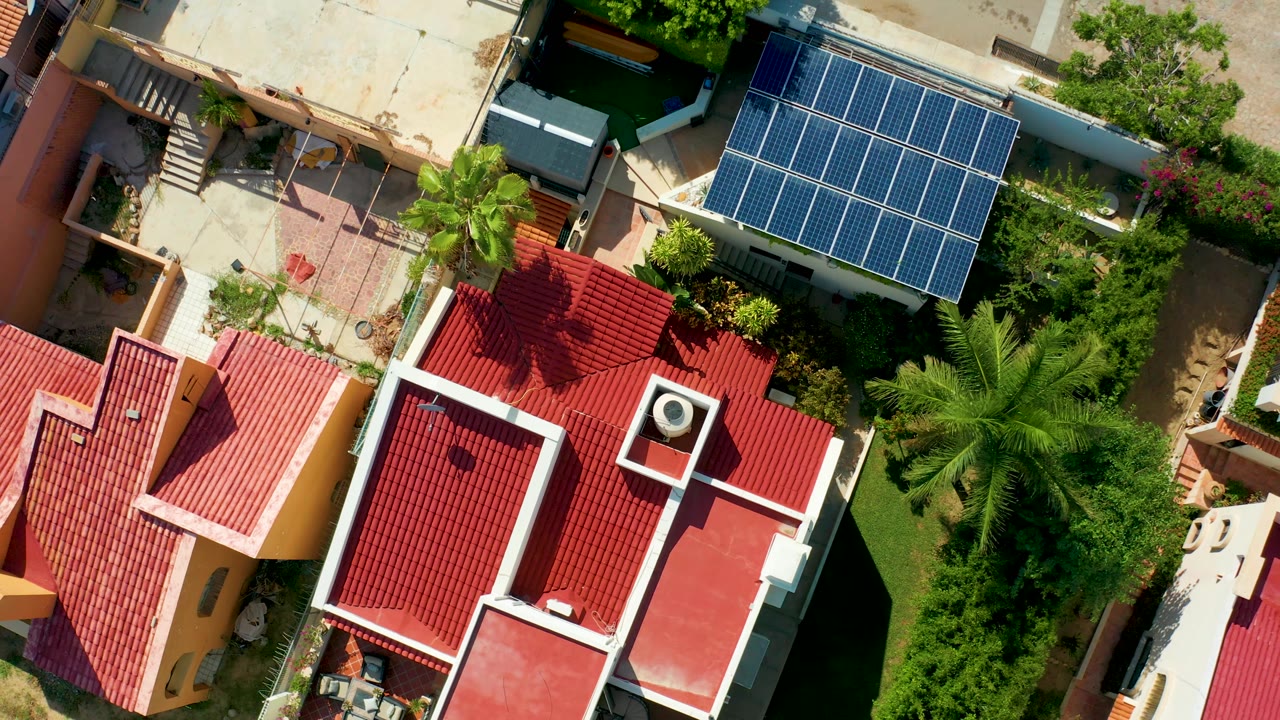 Aerial San Jose Del Cabo view showing red tile rooftops, white parapet lines, a large blue solar panel grid, palms, and green vegetation, with no visible people.