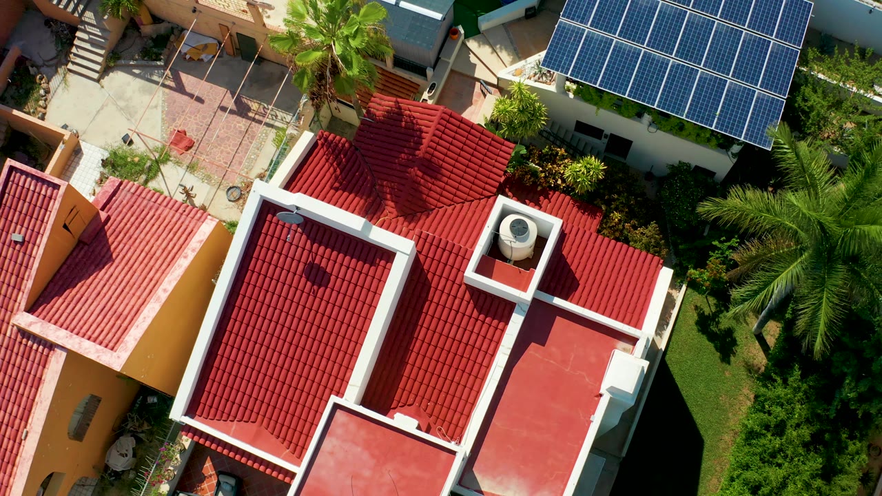 San Jose Del Cabo aerial frame with a red tile roof centered lower in the view, a pale courtyard and palm above it, blue solar panels at upper right, and no visible people.