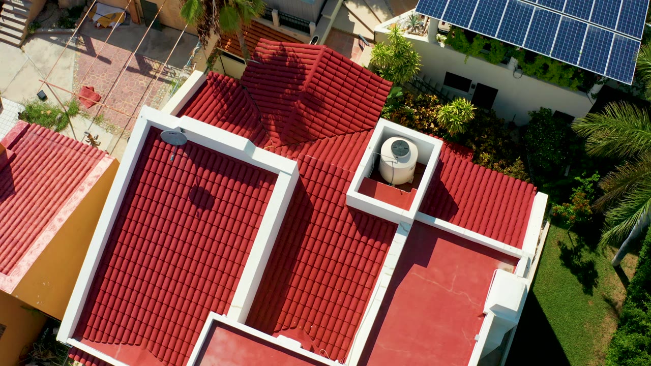 Overhead drone frame in San Jose del Cabo showing red clay roof tiles, white parapets, a rooftop water tank, courtyard paving, blue solar panels, palms, and no visible people.