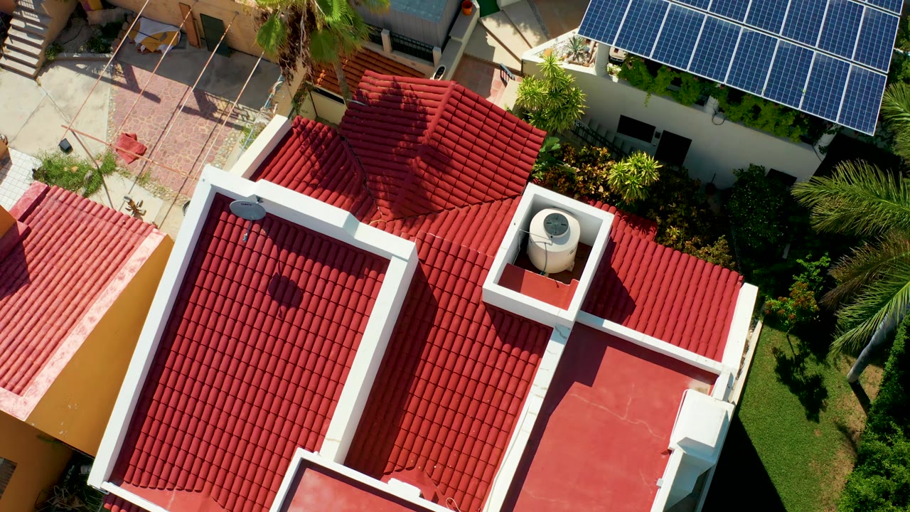 At 0:02, the San Jose del Cabo overhead frame shows the red tile roof mass, a flat red terrace, courtyard paving, a blue solar-panel grid, palms, dense greenery, and no visible people.
