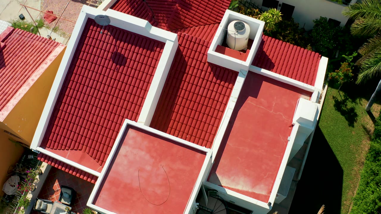 Aerial frame in San Jose Del Cabo with red clay roof tiles, a white-trimmed rooftop cylinder, flat red roof surfaces, garden greenery, and no visible people.