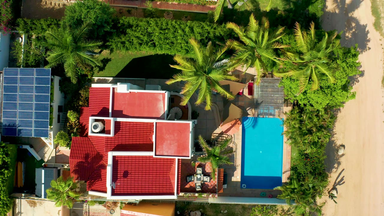 Overhead frame in San Jose del Cabo shows a red-tiled house beside a bright blue pool, palm trees, solar panels, sandy road, and no visible people.