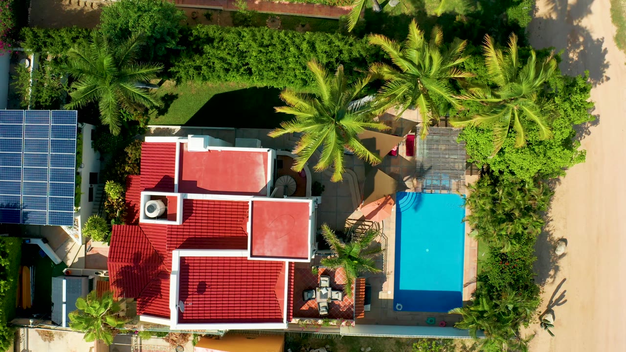 At 0:02, the overhead San Jose del Cabo view shows the rectangular blue pool on the right, red roof below center, solar panels at left, palms above, and no visible people.