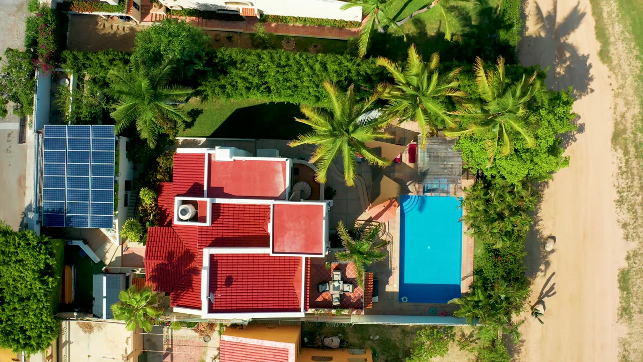 One second into the San Jose del Cabo overhead, the red roof sits lower in frame with the solar panels at left, the blue pool at right, dense greenery above, and no visible people.