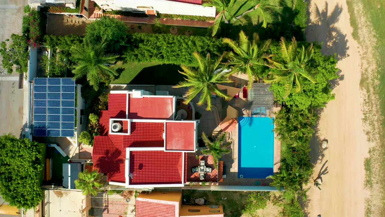 Overhead frame of a San Jose Del Cabo residential property with red roofs, solar panels at left, a blue pool at right, palms, greenery, sandy ground, and no clearly visible people.