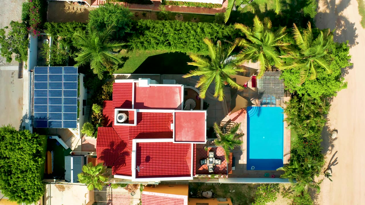 Overhead view of a residential lot with red roof, solar panels, palms, and blue pool cover.