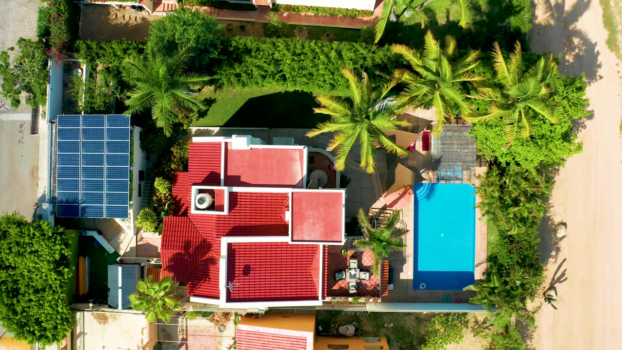 Top-down San Jose del Cabo view showing the house shifted slightly within frame, the blue covered pool along the right edge, a sandy road beyond the wall, solar panels to the left, and a small red-shirted person near the upper pool-side area.