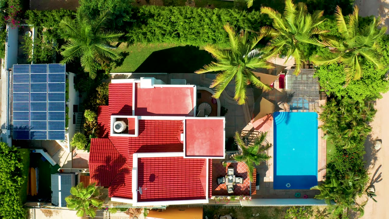 Top-down aerial frame in San Jose del Cabo showing a red-roofed residence, a bright blue rectangular pool at right, solar panels at left, dense greenery, patios, and a small person in the water.