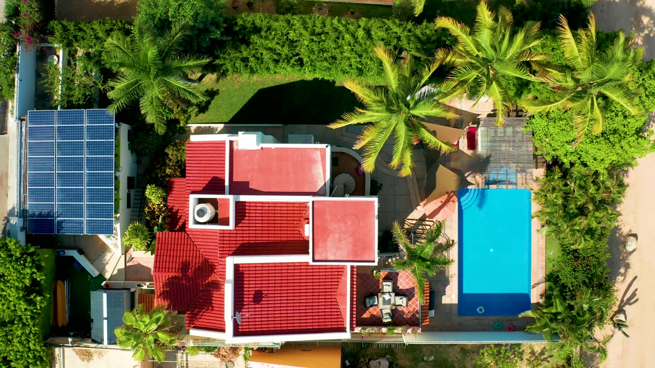 At 0:02, the overhead San Jose del Cabo frame shows the pool shifted slightly within the right side of the composition, with red roof planes, solar panels, palm shadows, and a small swimmer visible below.