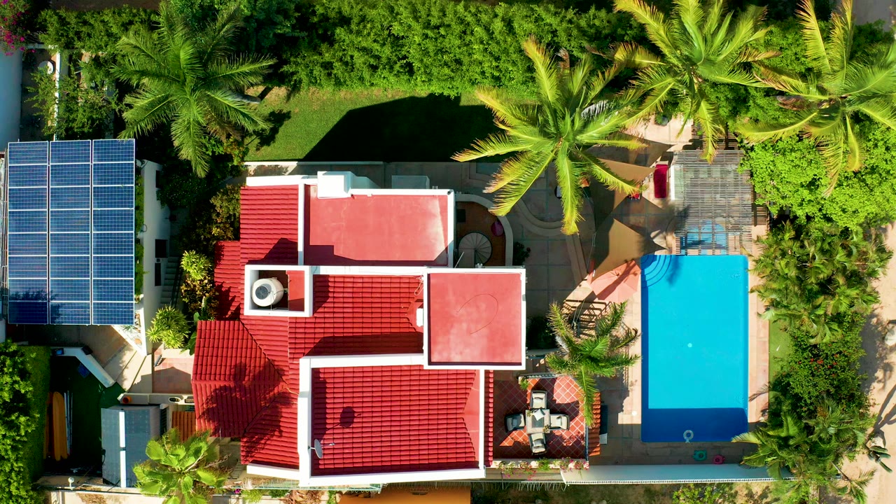 Opening overhead frame in San Jose del Cabo showing a red-roofed house, blue solar panels on the left, a turquoise pool on the right, palms, garden greenery, and no visible people.