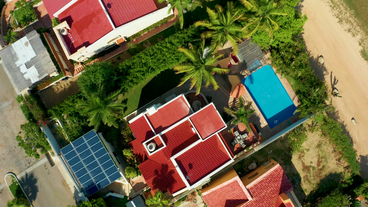 Top-down aerial frame in San Jose del Cabo showing red-roofed houses, dense palms, solar panels at lower left, a bright blue pool area at right, sandy road edges, and no visible people.