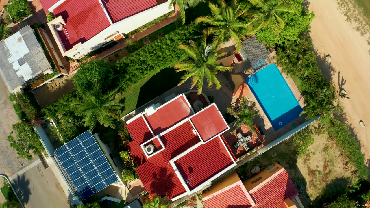 Overhead San Jose del Cabo frame showing red-tile rooftops near the top and center, thick green vegetation, a bright blue pool area to the right, sandy ground along the road, and no visible people.