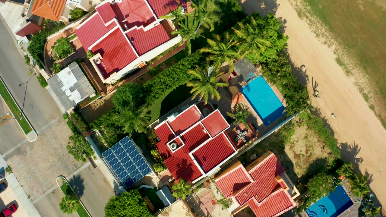 Top-down San Jose del Cabo frame with red roofs centered lower in view, a large solar-panel array near the street, palms around the houses, sandy shore to the right, and no visible people.