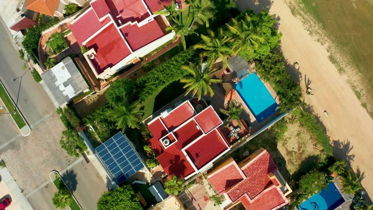 Aerial San Jose del Cabo view with the red-roofed houses shifted slightly lower, palms filling the center, beach sand along the right edge, a paved street at left, and no visible people.