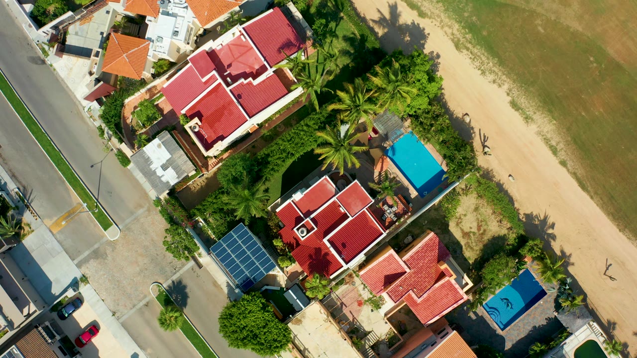 Aerial neighborhood view with red roofs, blue pool, palms, street, and sandy strip.