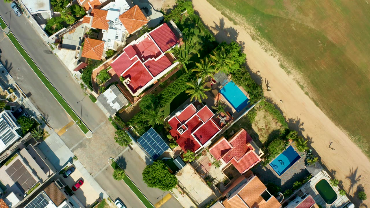 Overhead frame in San Jose Del Cabo showing red-roofed homes, a paved street, blue pools, palm trees, sandy beach, shallow water, and no visible people at the opening moment.