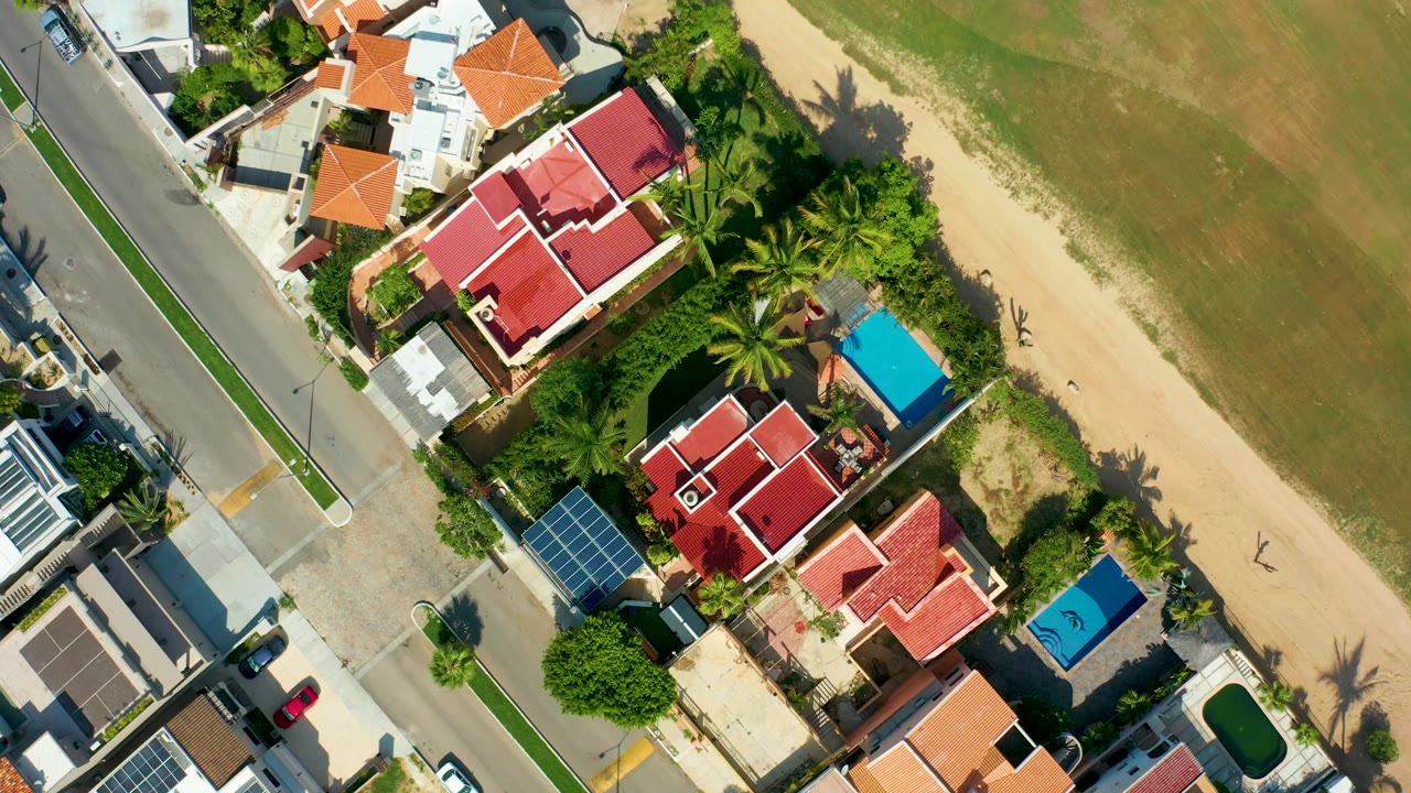 One second into the San Jose Del Cabo aerial, the road and red-roofed houses remain on the left while the sandy beach and green-brown water fill the right, with no visible people.