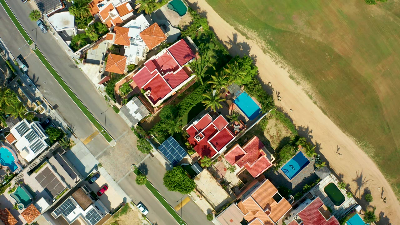 Overhead aerial frame in San Jose Del Cabo showing a residential road, red-roof houses, palms, pools, a sandy path, and a green field with no visible people.