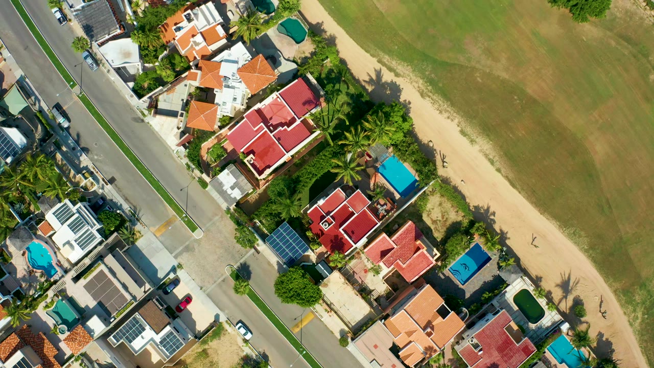 Overhead view in San Jose Del Cabo, Mexico showing a coastal road, red-roofed houses, blue pools, palm trees, a sandy strip, and grass with no visible people.