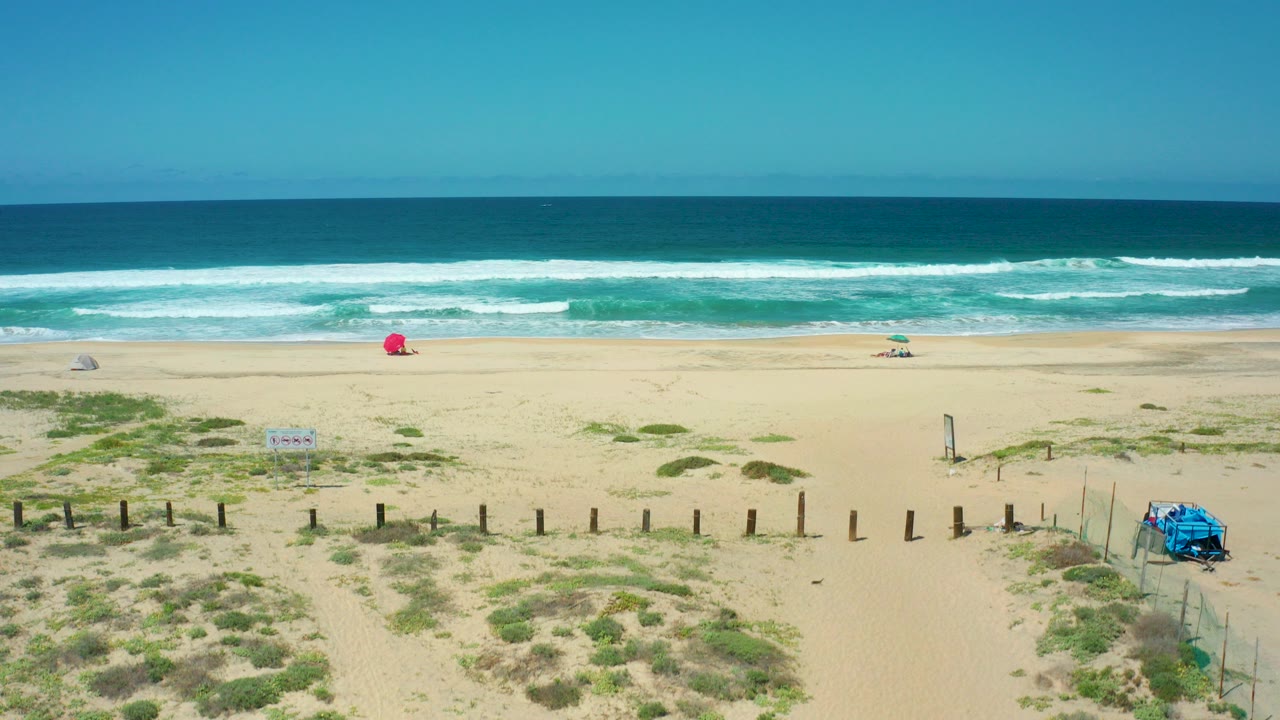 Wide aerial view of beach dunes, surf, clear sky, red umbrella, and blue beach structure.