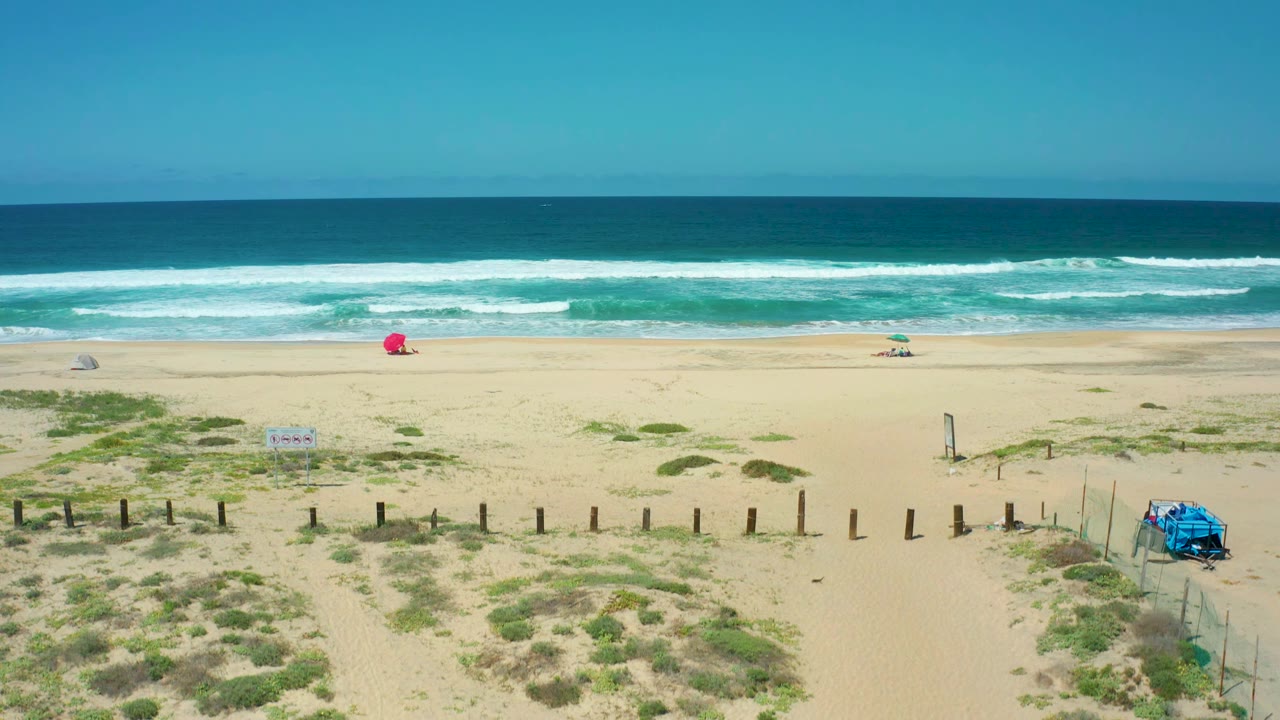 At 0:01, the aerial view over Cerritos Beach shows more dune foreground, a sandy path beside fence posts, breaking ocean waves, the red umbrella in the distance, the blue structure at right, and no visible people.