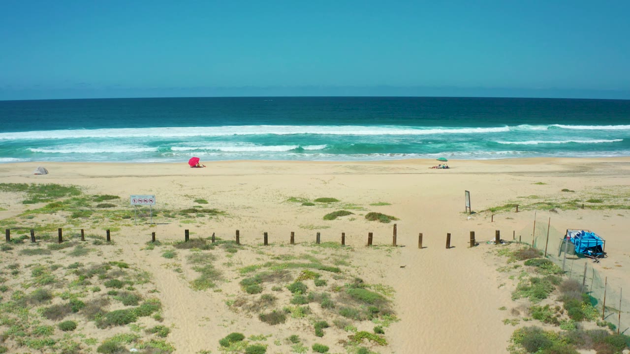 At 0:02, Cerritos Beach is framed with sandy paths running through low green shrubs, fence posts across the midground, layered surf in the background, the red umbrella left of center, the blue structure at right, and no visible people.