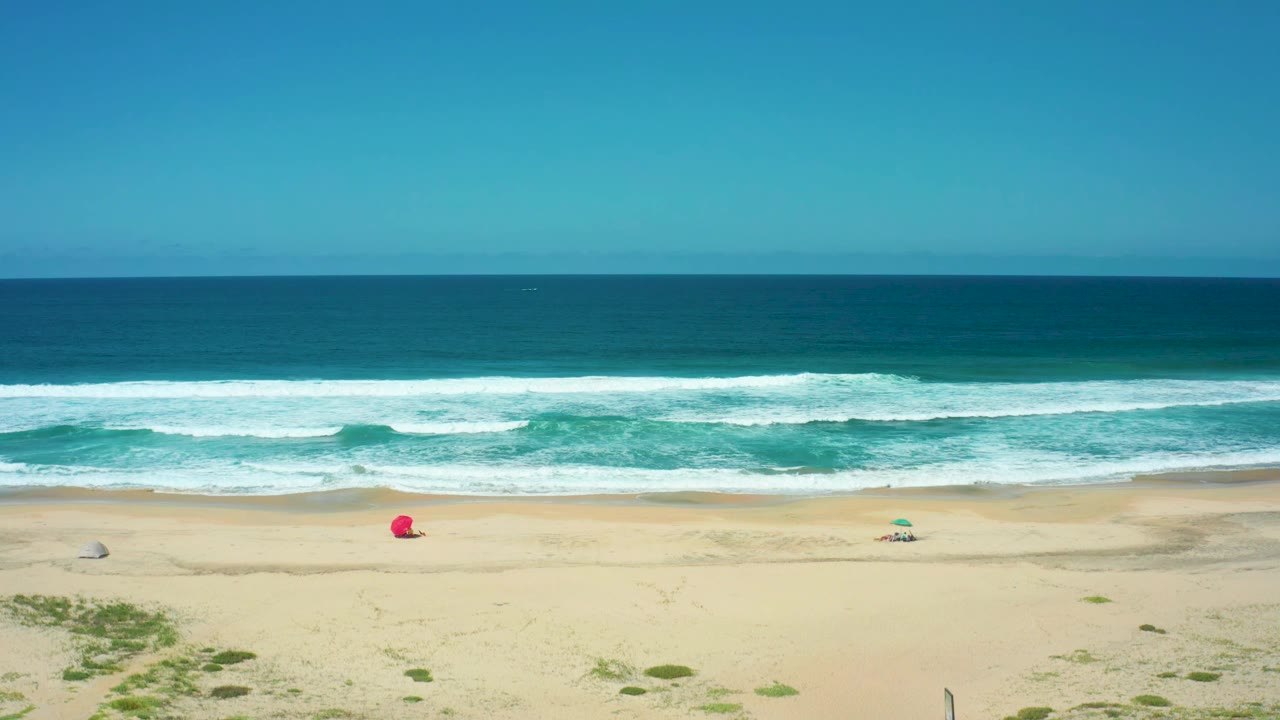 Aerial view over Cerritos Beach showing layered whitecaps, a broad empty sand strip, a red object near the beach center, small umbrellas, and no visible people.