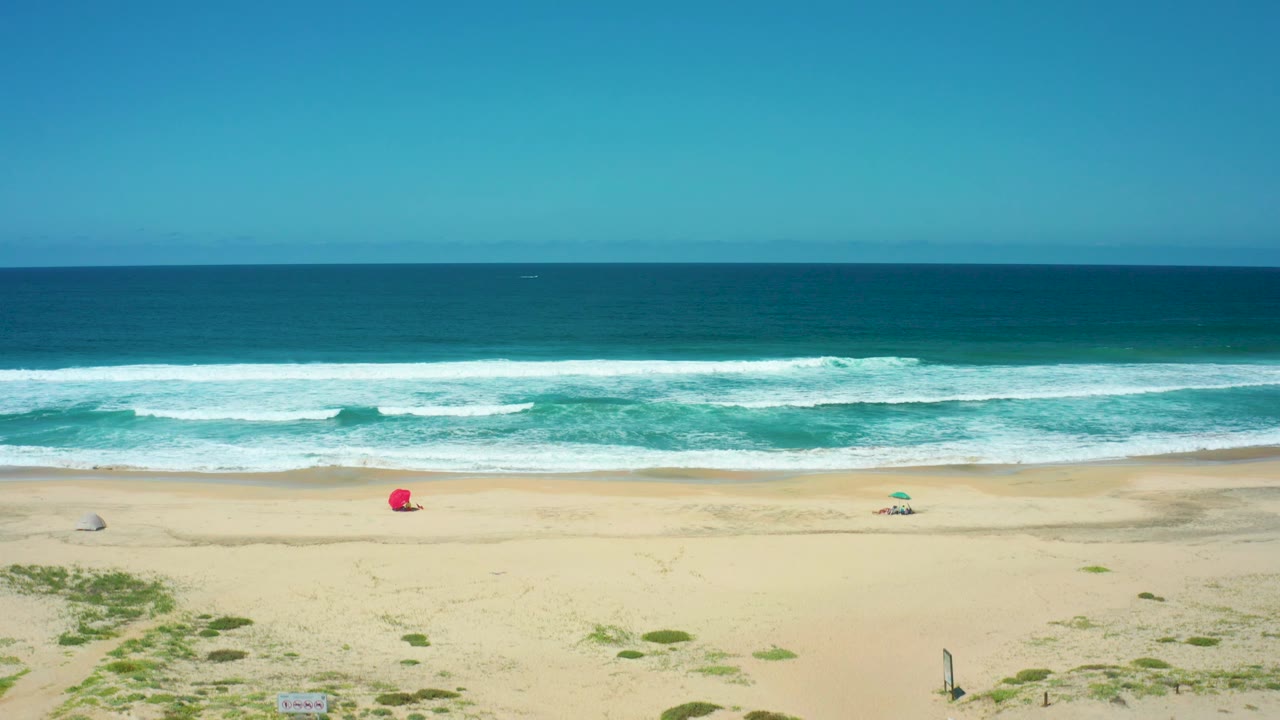Cerritos Beach appears from a high coastal angle with teal surf, long white wave lines, pale sand, a red object, scattered small umbrellas, dune greenery, and no visible people.